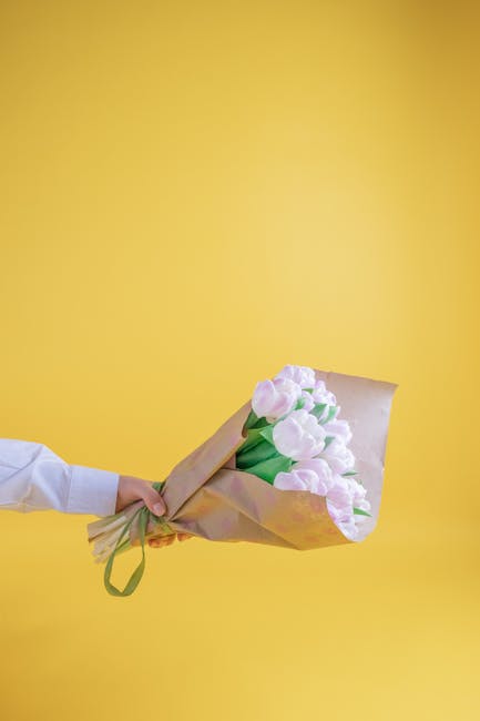 A person in a white shirt holds a bouquet of fresh pink tulips wrapped in light brown paper and tied with a green ribbon, presented against a vibrant yellow background. The tulips have slightly open p