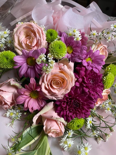 A vibrant floral arrangement featuring soft pink roses with fully bloomed, gently curled petals, surrounded by purple daisies with yellow centers, bright green pom-pom chrysanthemums, and small white