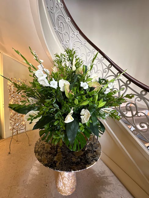 A large, elegant floral arrangement featuring white lilies, anthuriums, and various lush green foliage, including broad leaves and delicate stems, arranged in a black vase. The bouquet is placed on a 