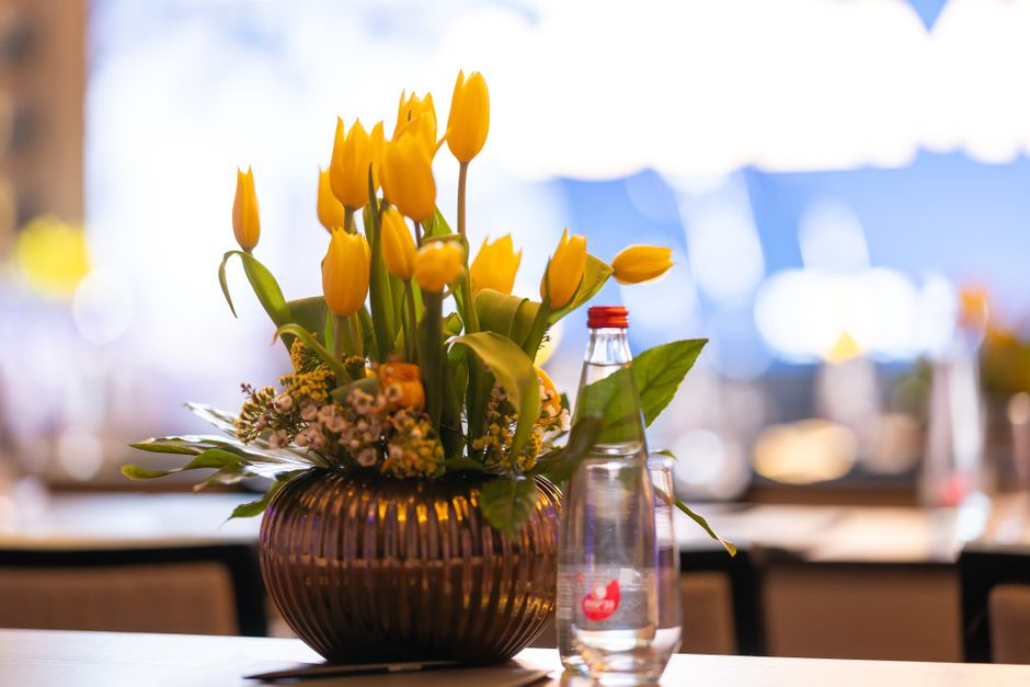 A floral arrangement featuring tall, vibrant yellow tulips with closed buds and lush green leaves, set in a round, brownish-tinted glass vase with vertical ridges. The bouquet includes small pink and 