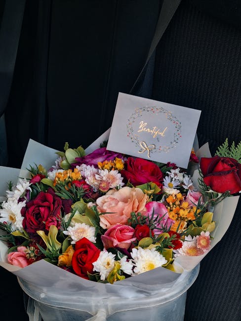 A vibrant bouquet of fresh flowers arranged in a generous, circular bouquet features red roses, pink roses, peach lilies, yellow and white chrysanthemums, and small white daisies, all wrapped in trans