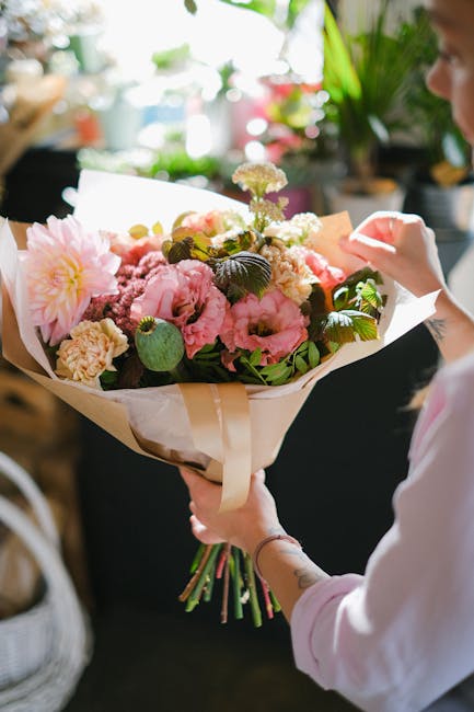 A person holding a vibrant floral bouquet arranged with pink peonies, soft white hydrangeas, and greenery, wrapped in cream-colored paper with a satin ribbon. The bouquet is being displayed indoors ne